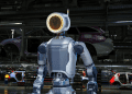 A sleek, electric Atlas robot with a ring-shaped head unit stands in a factory setting, poised to perform a task.