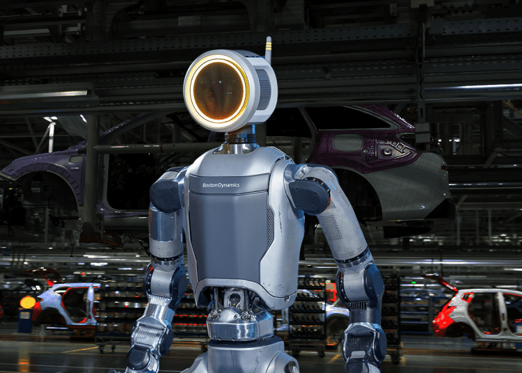 A sleek, electric Atlas robot with a ring-shaped head unit stands in a factory setting, poised to perform a task.