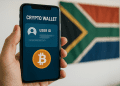 Smartphone showing crypto wallet screen with Bitcoin icon and blurred user ID, held in front of a South African flag — FATF Travel Rule context.