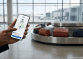 A person using an Android phone to share a luggage tracker's real-time location via Google Find Hub at an airport baggage carousel.