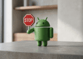 A 3D minimalist figurine of the green Android robot holding a small red stop sign, standing on a grey concrete surface with a blurred interior background.