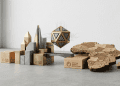 A minimalistic 3D editorial design featuring a stylized geometric crypto token levitating between a modern brass and stone skyline and a wooden map of Africa, representing GenZ driving the next crypto revolution.