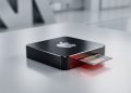 A minimalistic, 3D photograph of a sleek, dark-brushed metal Apple hardware component centered on a clean silver surface, with a single microchip protruding from a slot illuminated by a subtle red warning light.