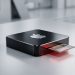 A minimalistic, 3D photograph of a sleek, dark-brushed metal Apple hardware component centered on a clean silver surface, with a single microchip protruding from a slot illuminated by a subtle red warning light.