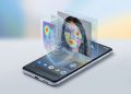 A 3D-style editorial illustration of an Android smartphone on a minimalist background. Holographic layers rise from the screen, showing an analytical wireframe, a facial recognition heatmap overlaying a person's face, and a digital security shield, symbolising Google's real-time deepfake detection technology.