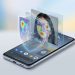 A 3D-style editorial illustration of an Android smartphone on a minimalist background. Holographic layers rise from the screen, showing an analytical wireframe, a facial recognition heatmap overlaying a person's face, and a digital security shield, symbolising Google's real-time deepfake detection technology.