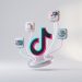 A minimal, 3D editorial graphic showing the TikTok logo at the centre, connected by glowing neon lines to icons for shopping, banking, video messaging, and global networking against a clean, light grey background.