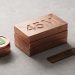 A minimal editorial 3D photograph showing a stack of metallic copper blocks engraved with '45M' alongside a stack of coins featuring the green Cash App logo and a brass ruler engraved with 'STATE PROBE' on a clean concrete background.